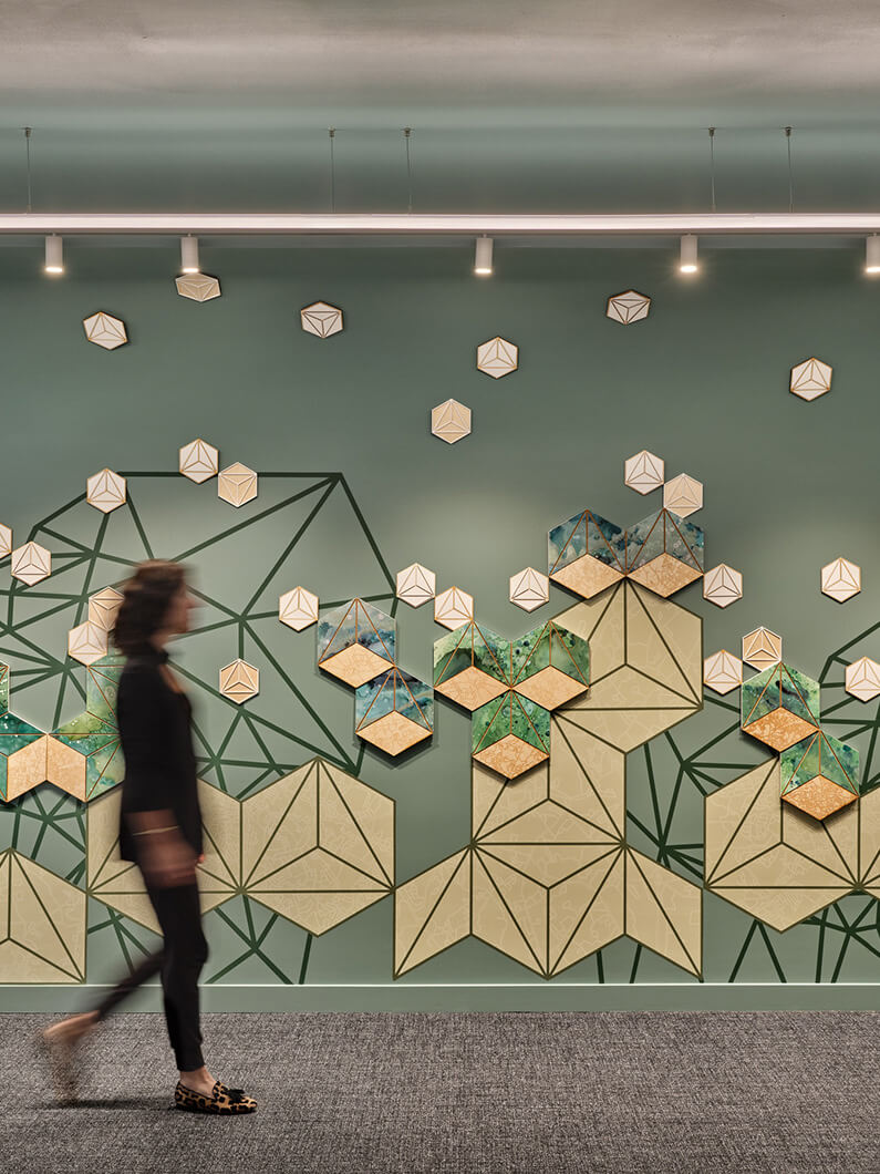 A person walks past a geometric wall installation with green and beige shapes, featuring light fixtures above.