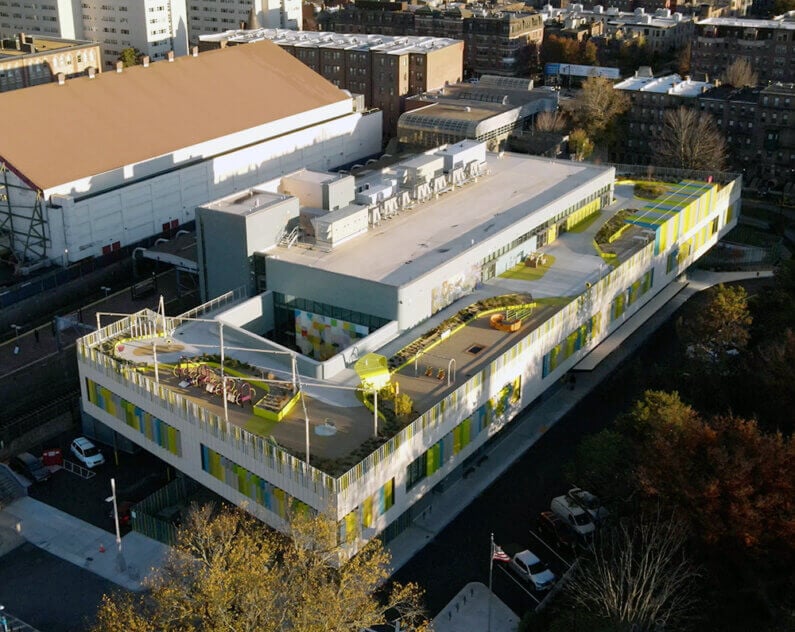 Aerial view of a green-roofed school building with play areas and gardens, surrounded by urban architecture and tree canopies.