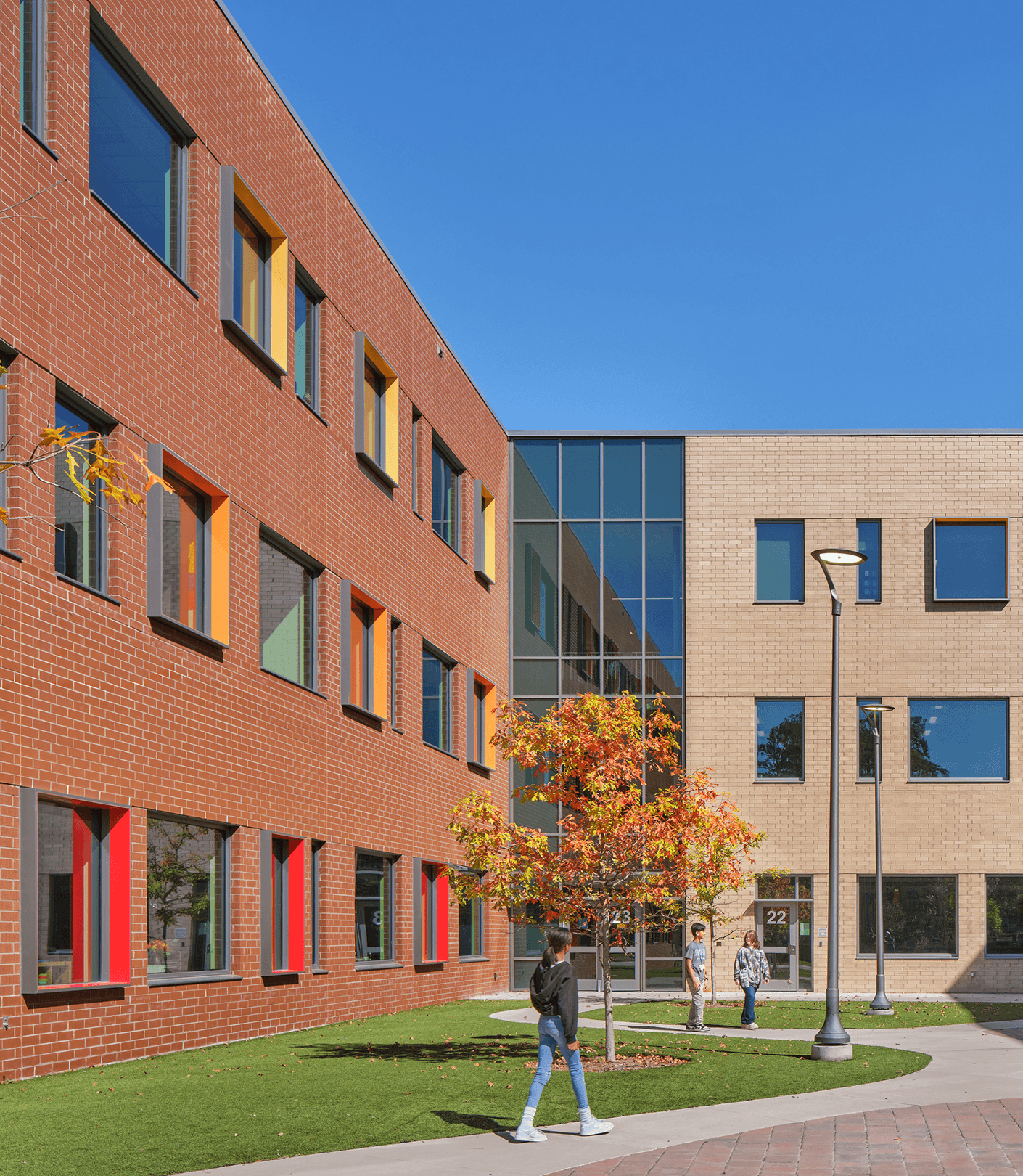 Two people walk in an outdoor area with a red-brick building and glass facade. Colorful windows and a small tree with autumn leaves are visible.