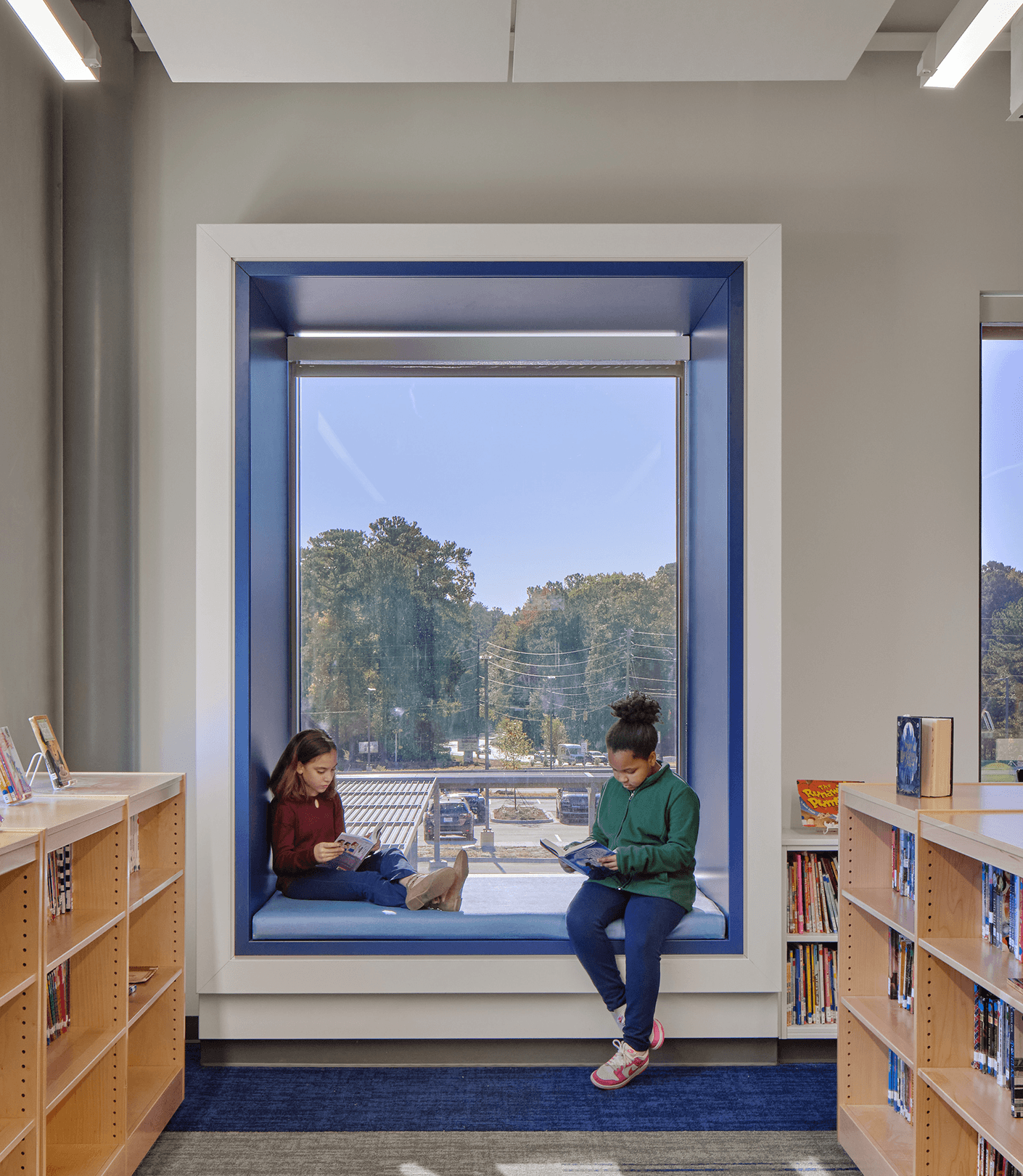 Two children reading in a window seat at a library, surrounded by shelves and natural light, with a view of trees and cars outside.