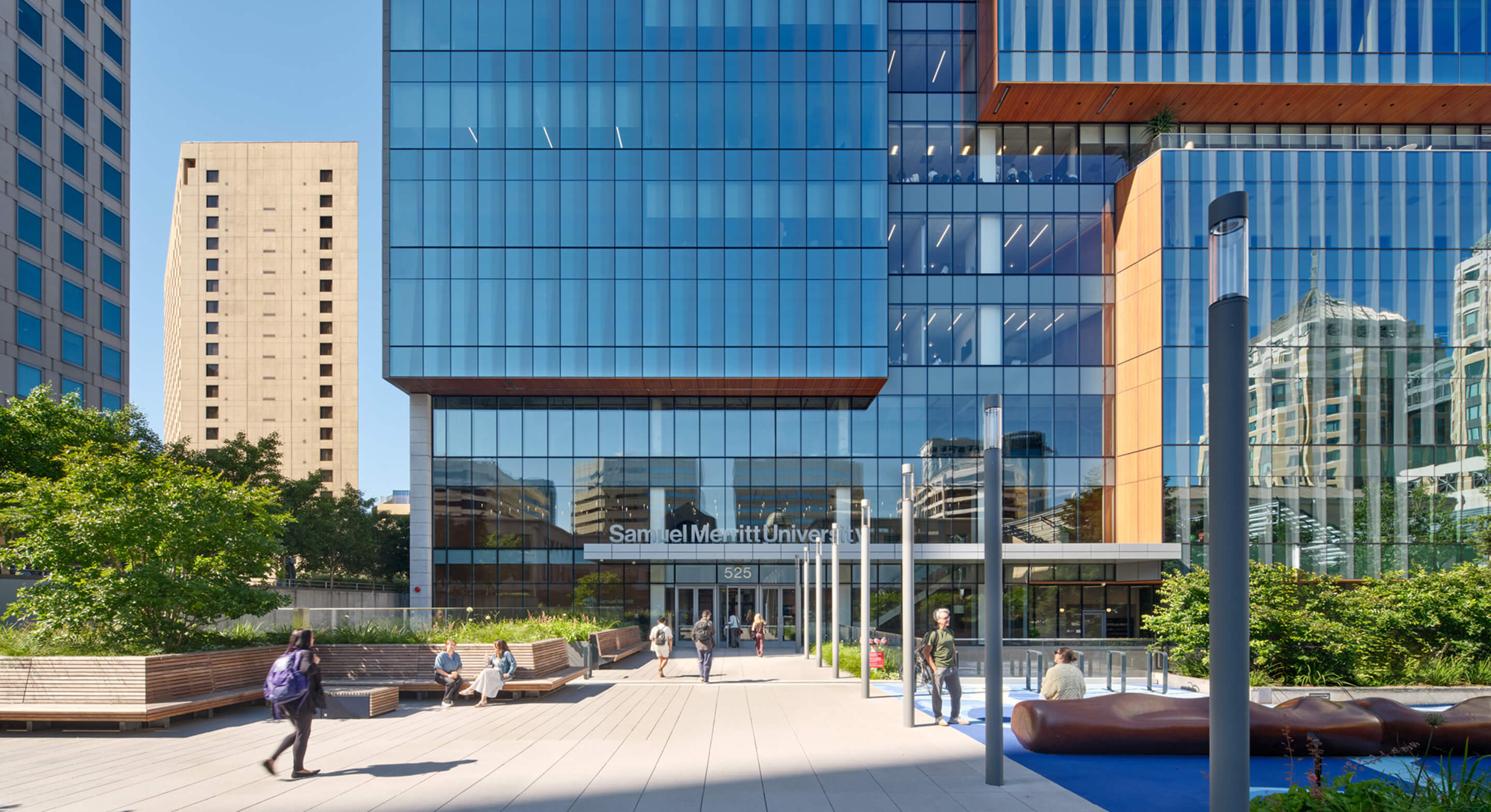 Students walk in front of the glass facade of Samuel Merritt University, with green landscaping and urban buildings in the background.