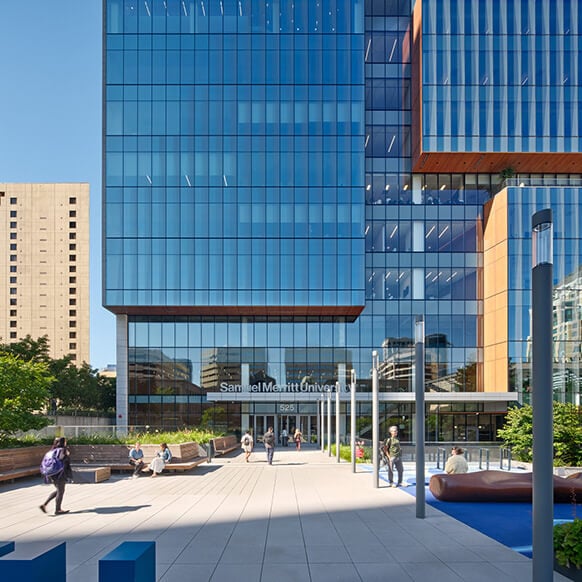 Facade of Samuel Merritt University building with glass exterior, landscaped area, and people walking in front on a sunny day.