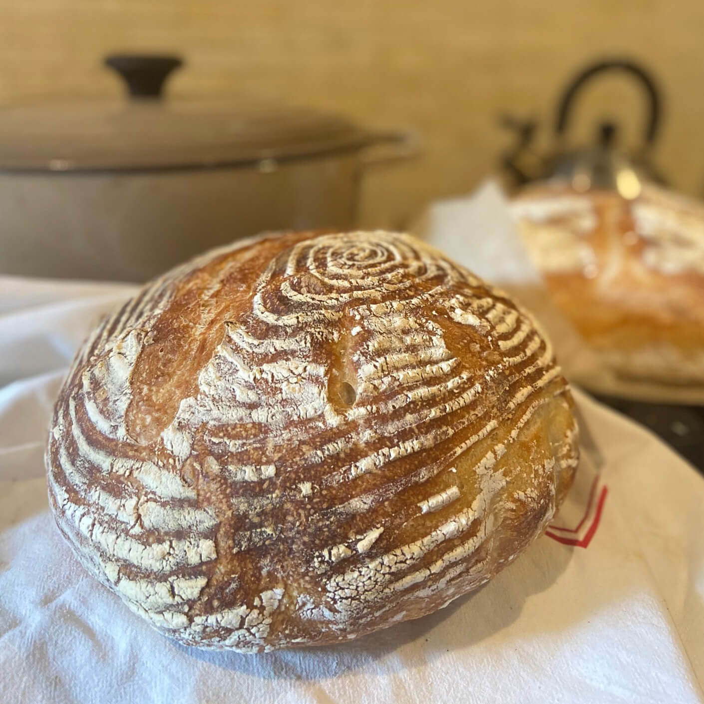 Artisan bread with a crusty surface, resting on a cloth, with a pot in the background. Two loaves of bread are visible.