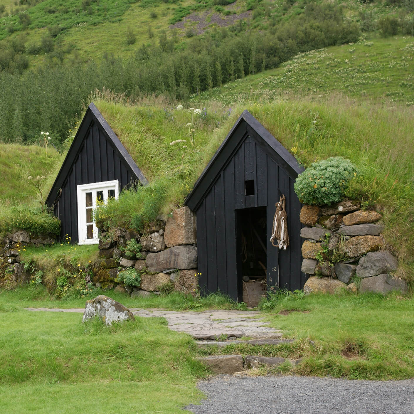 Black wooden houses with grass roofs nestled in a hillside, surrounded by lush greenery and stone walls. Pathway leading to entrance visible.