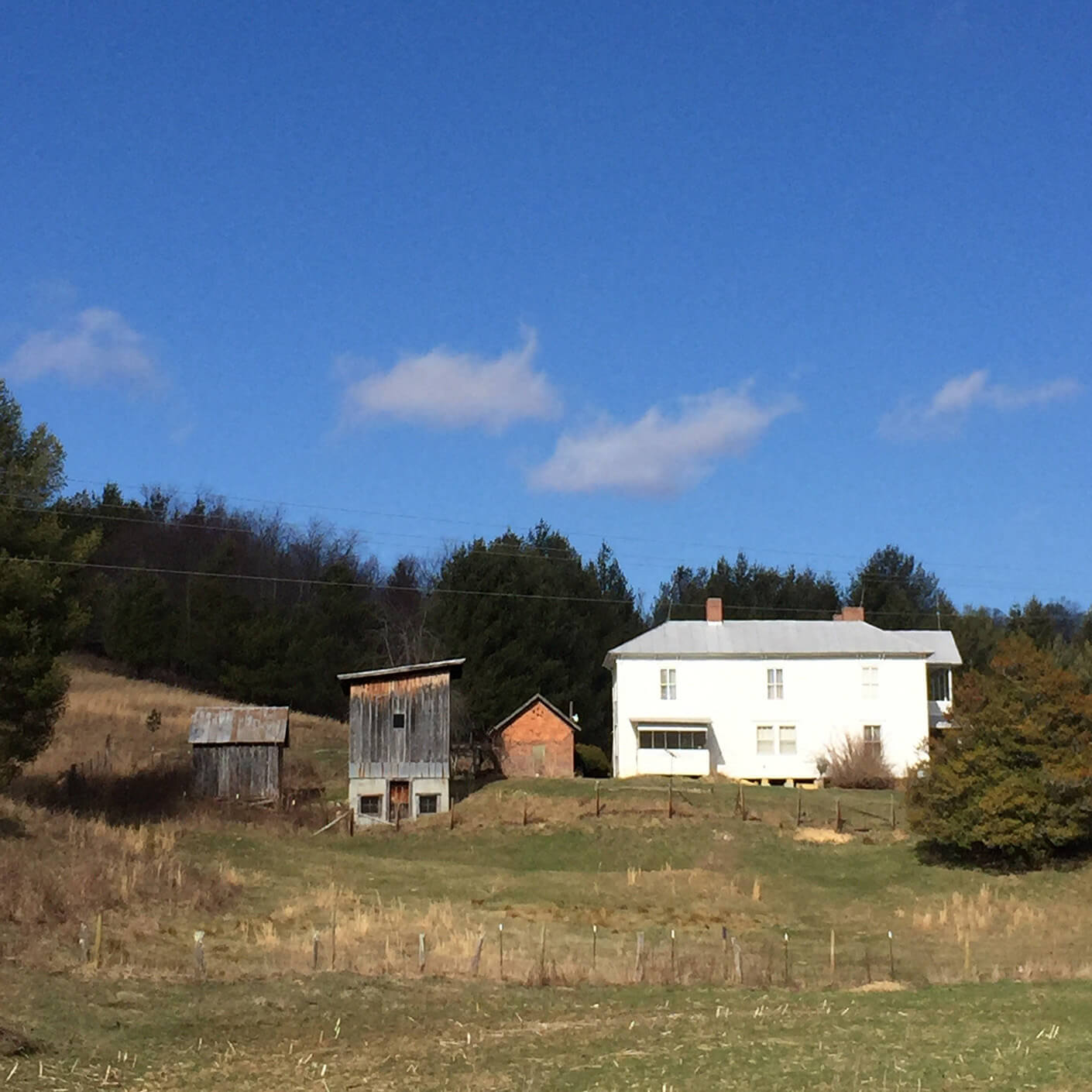 Historic farmhouse and outbuildings on grassy land, surrounded by trees and a clear blue sky. Rural landscape with no people.