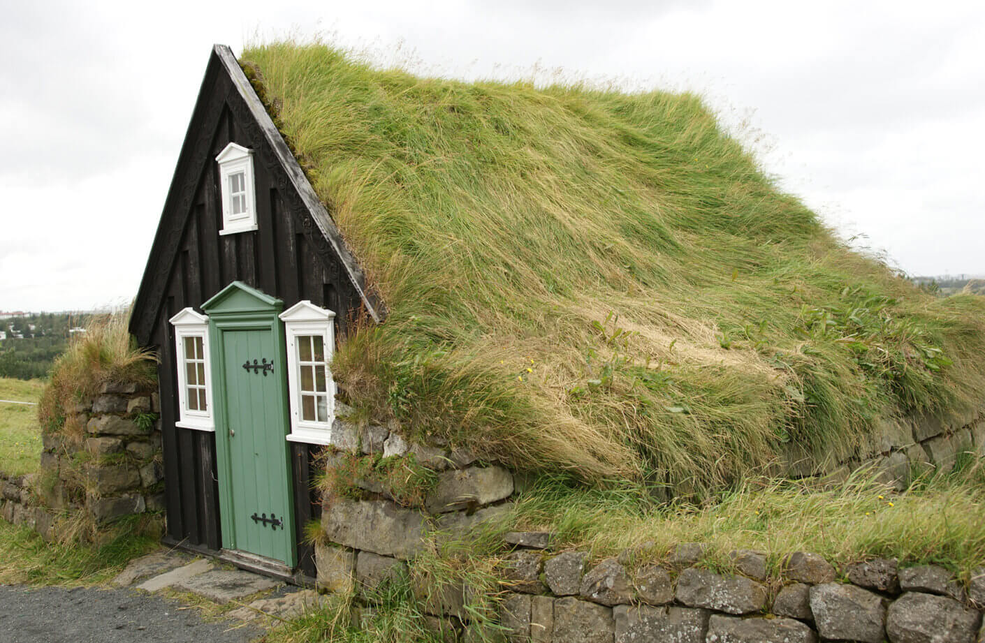 Grass-covered roof of a traditional cottage with a green door and white windows, surrounded by stone walls and open landscape.