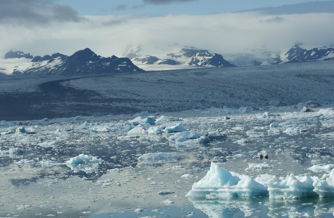 Icelandic glacial landscape featuring icebergs floating in a calm glacial lagoon, surrounded by mountains and a cloudy sky.