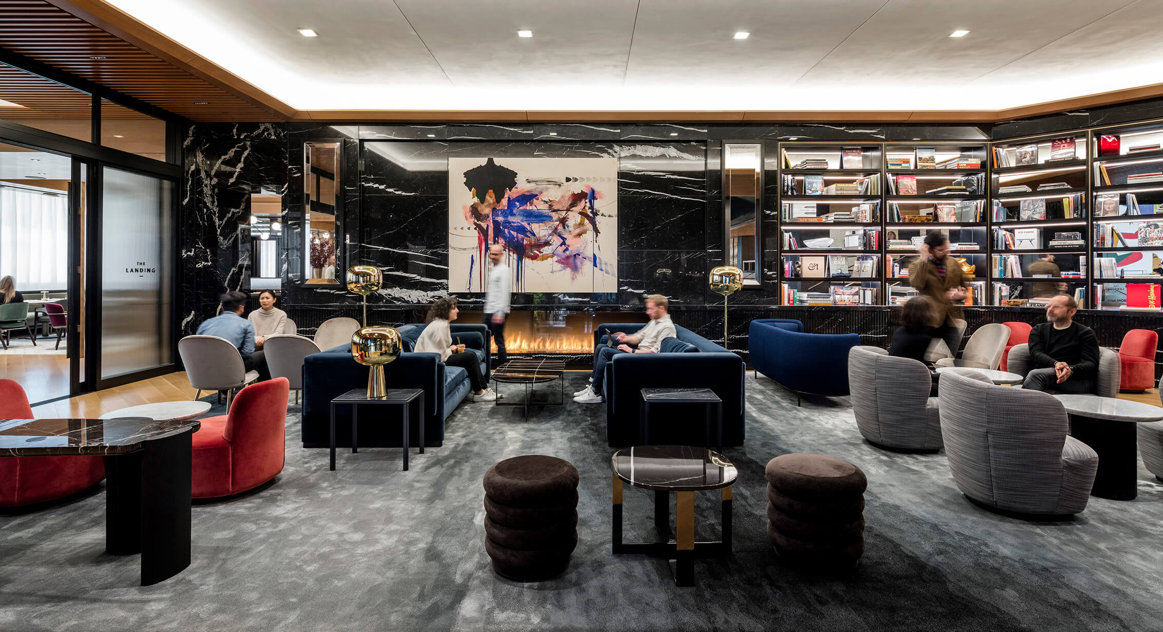 Interior lounge area featuring contemporary seating, a bookshelf, artwork, and a fireplace, with people engaged in conversation and relaxation.
