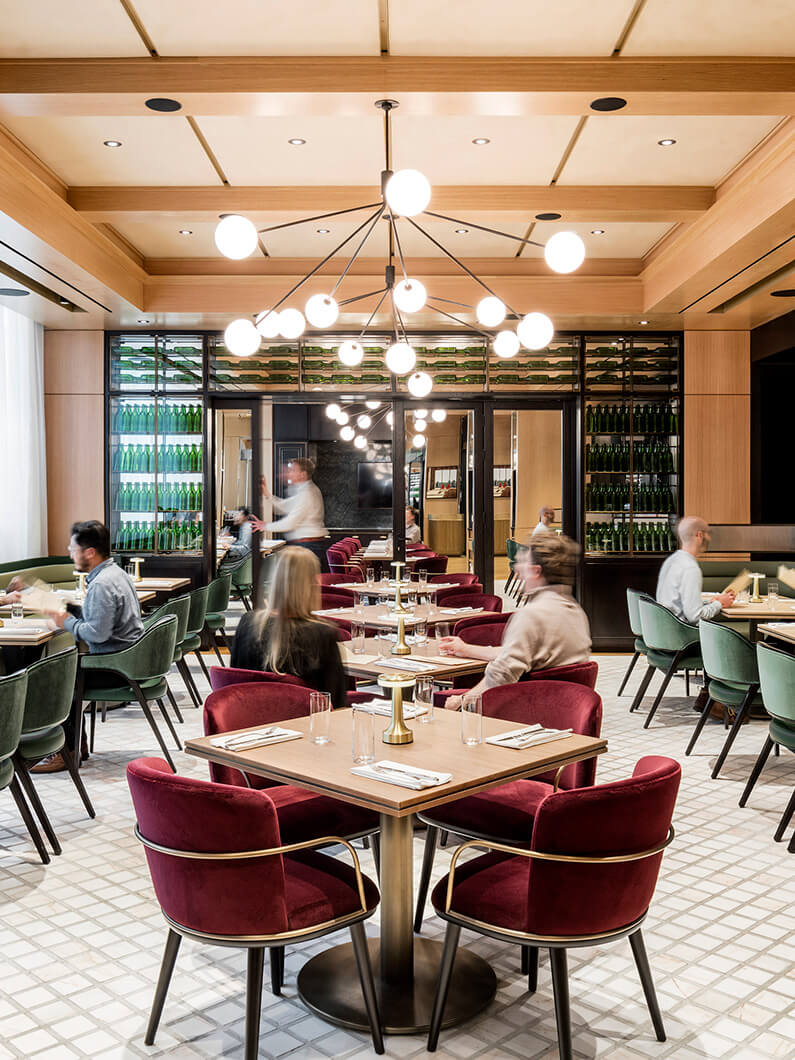 Interior view of a restaurant featuring round tables, plush chairs, and a pendant light fixture, with patrons dining and a blurred staff member serving.