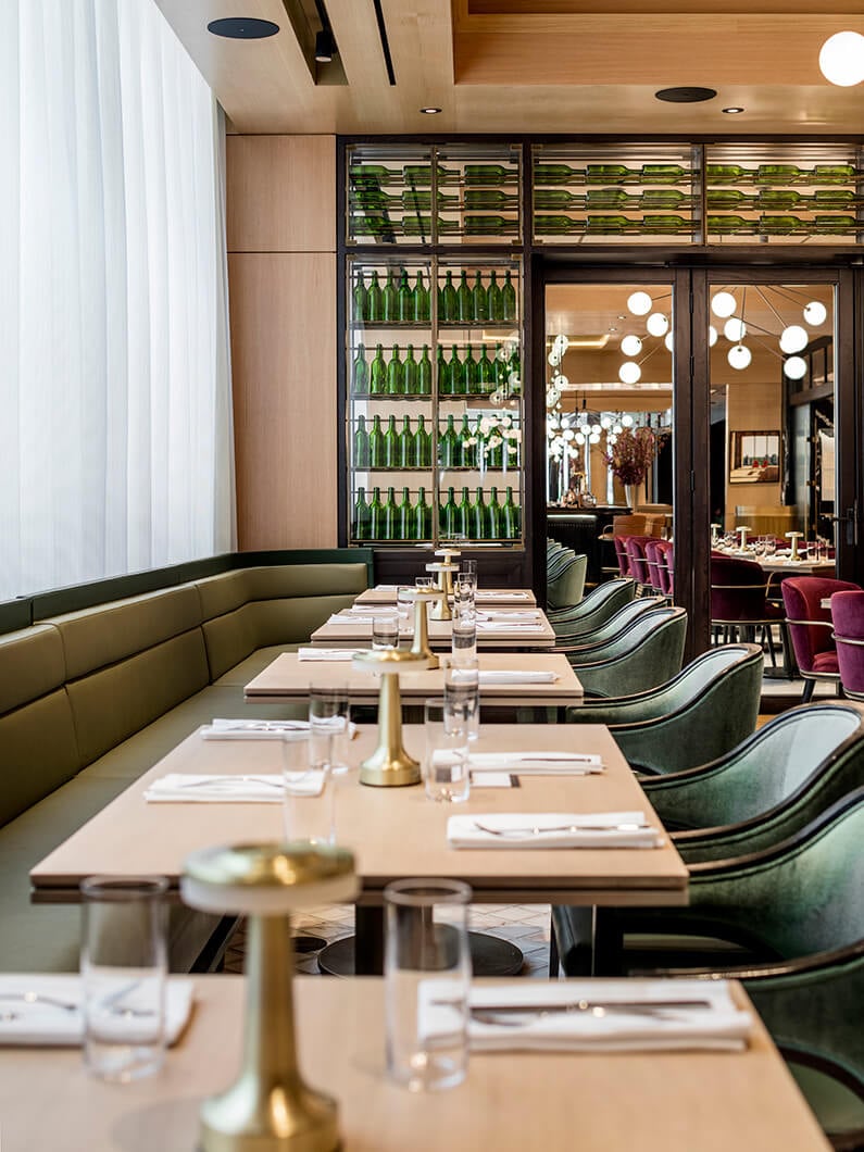Interior of a restaurant featuring green seating, wooden tables, glassware, and a wall of green bottles. Soft lighting enhances the dining area.