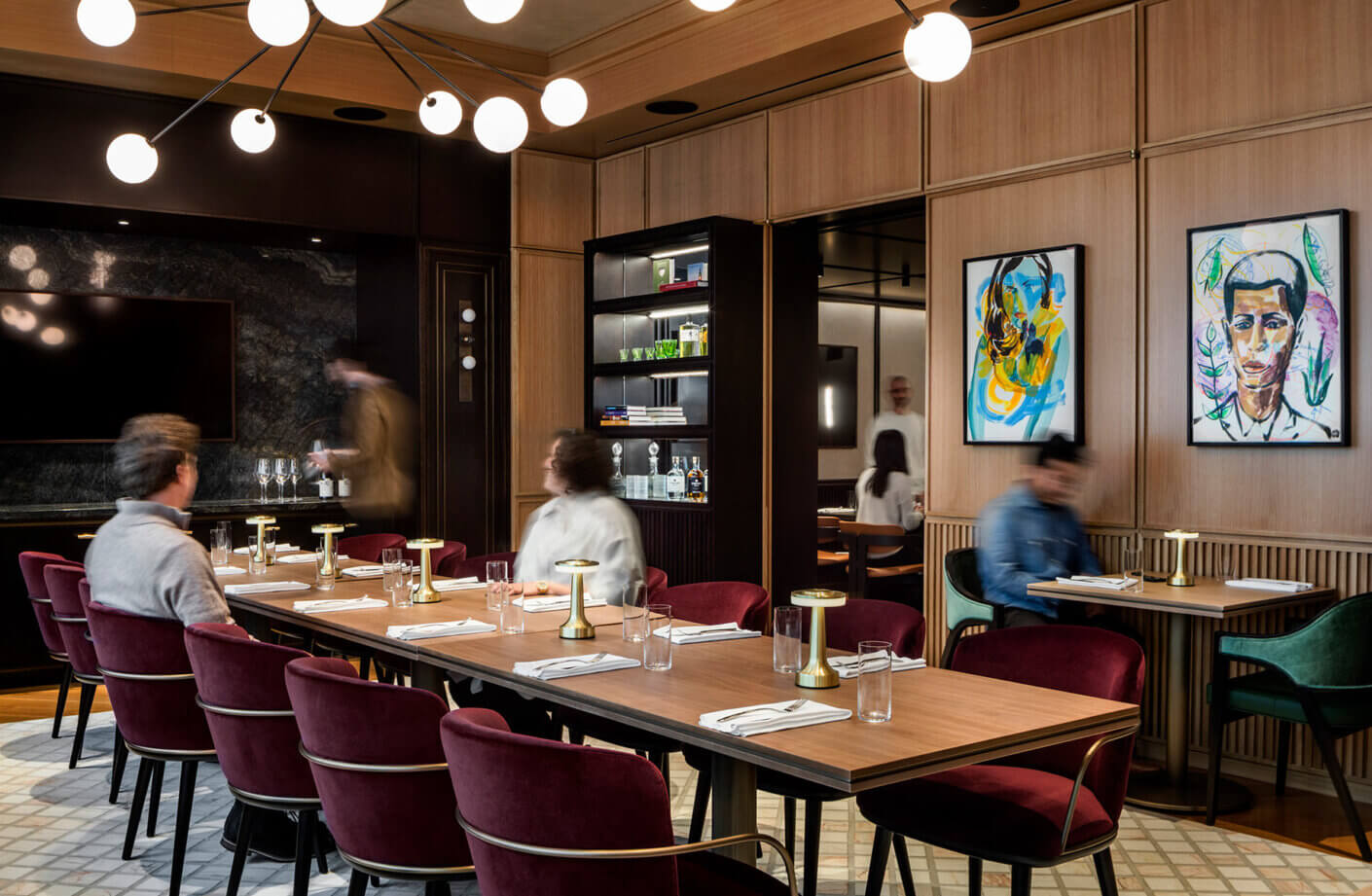 Interior view of a stylish restaurant featuring a long wooden table, plush chairs, and artistic wall decor, with guests enjoying their meals.