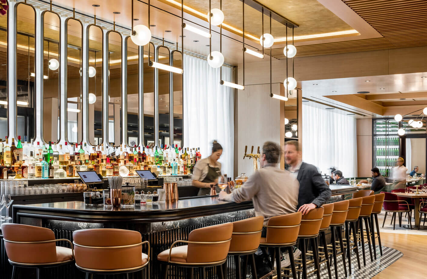 Interior of a stylish bar featuring a polished counter, various liquor bottles, and patrons engaging with staff. Bright lighting enhances the ambiance.