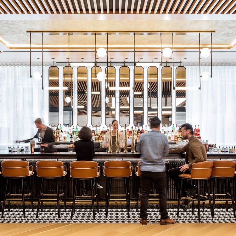 Bar scene with patrons and staff. A bartender prepares drinks behind a stylish counter. Sleek seating and decorative lighting enhance the space.