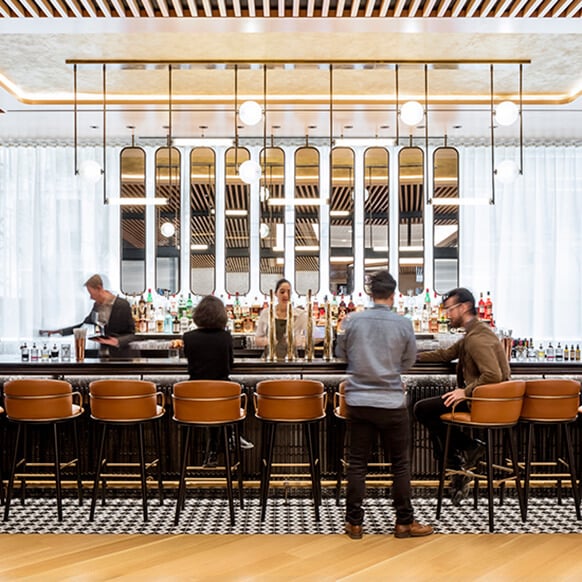 Bar scene with patrons seated at a stylish counter, a bartender serving drinks, and a diverse selection of bottles on display in a well-lit space.