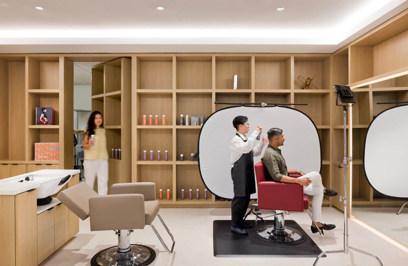 Hair salon interior with a stylist cutting hair while a client sits in a red chair; shelves display hair products and supplies.