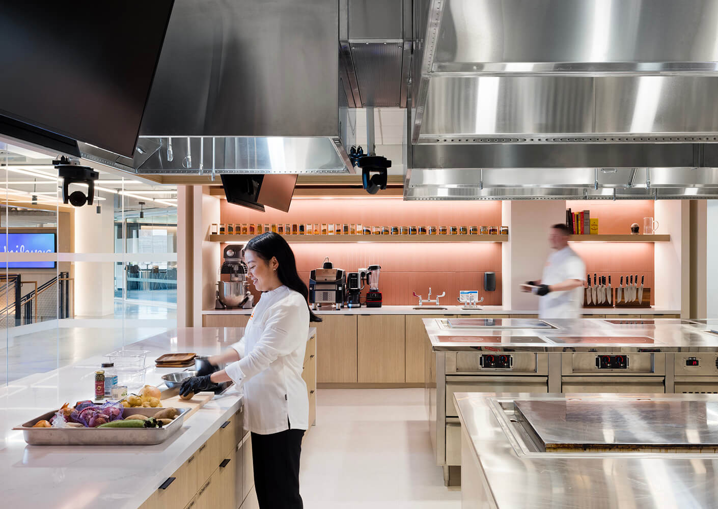 A chef in a kitchen preparing ingredients on a counter with stainless steel appliances and a blurred figure in the background.