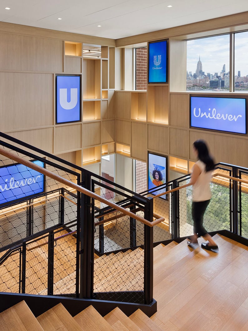 Staircase in Unilever office featuring wood accents, framed screens, and a view of the New York City skyline. Person walking down the stairs.
