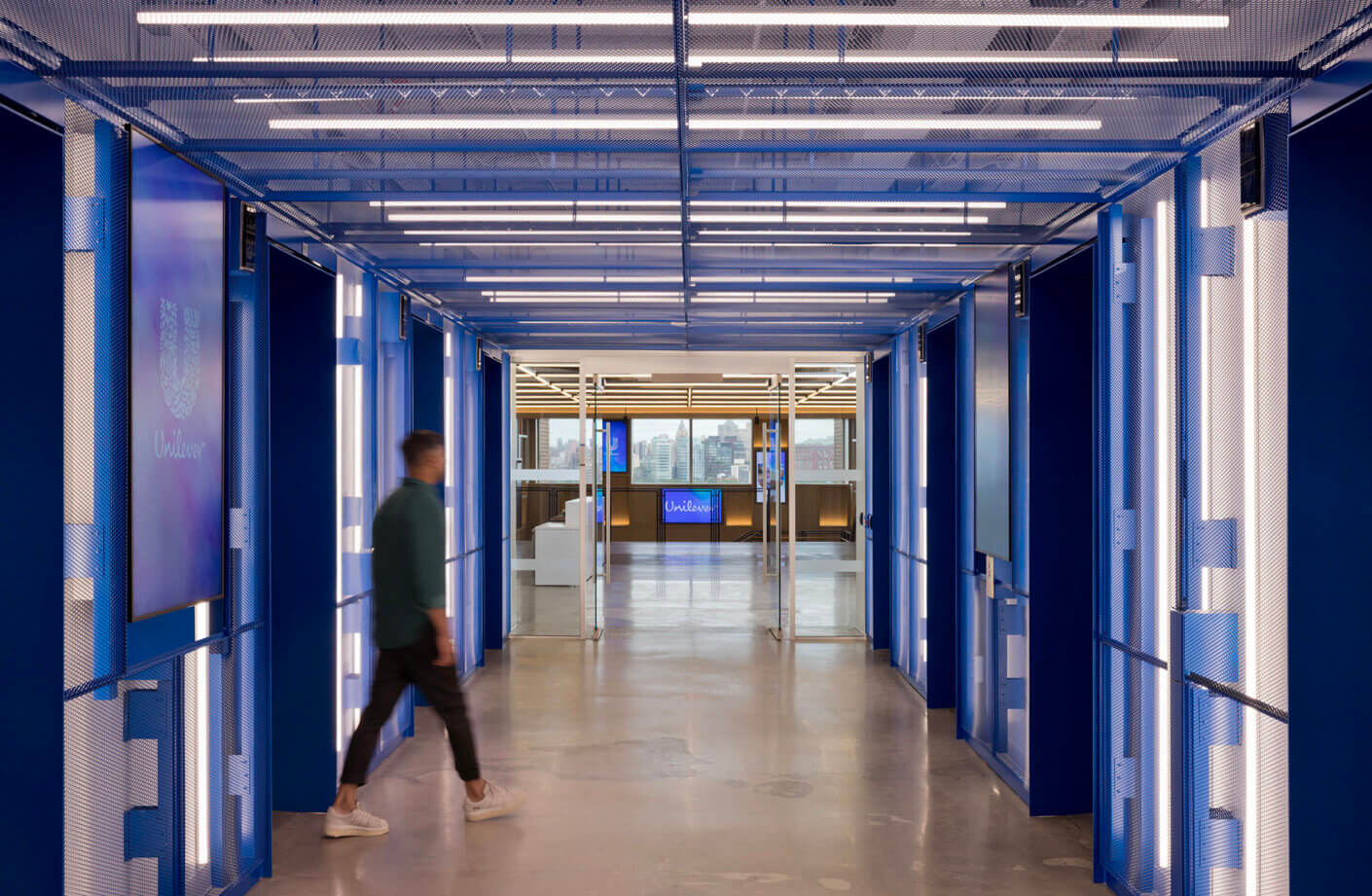 Bluish corridor with hanging lights and screens, featuring a person walking towards a reception area at Unilever's office.