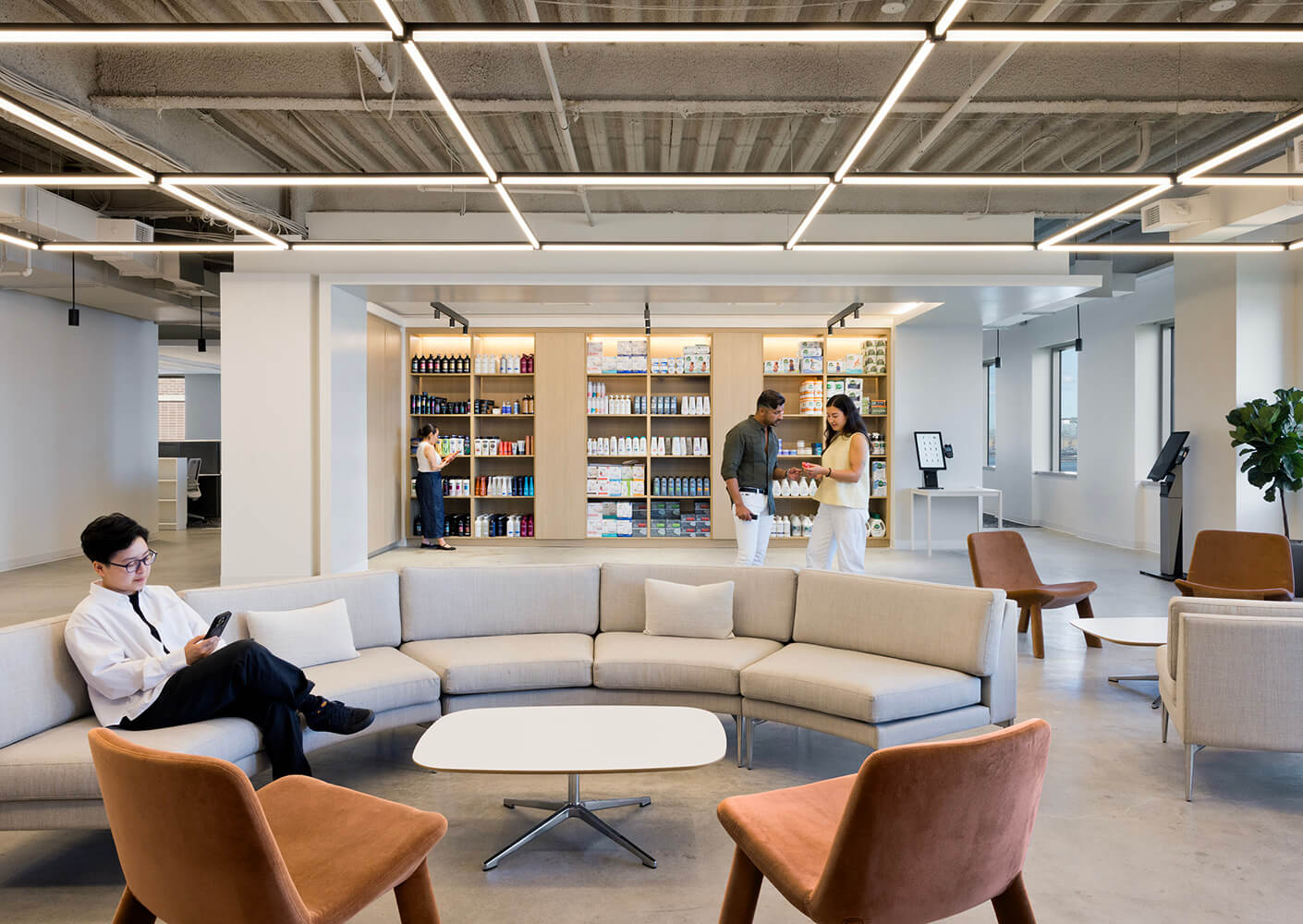 Office interior with a seating area, people interacting, and a wall display of products. Bright lighting and open layout evident.