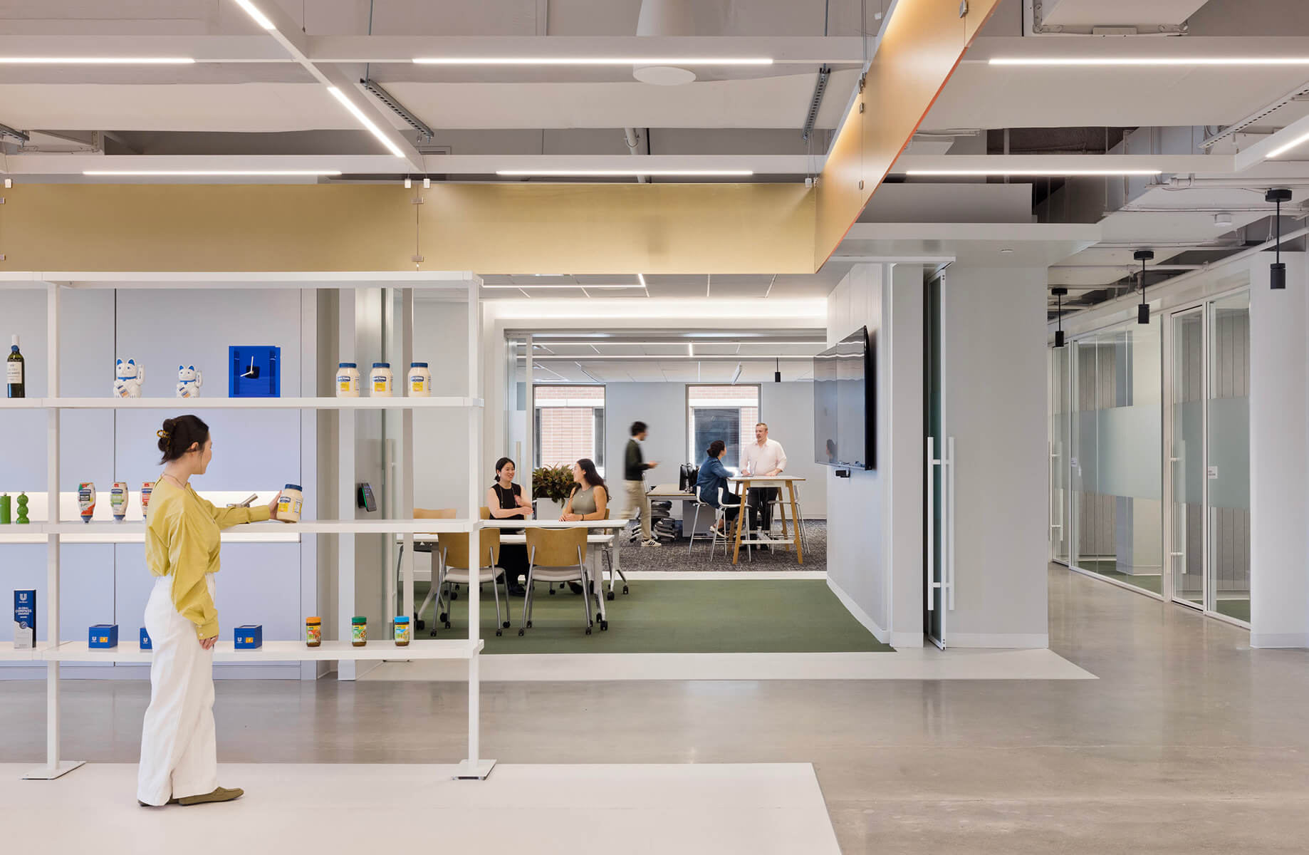 Office interior showcasing a woman examining products on a shelf, with a group collaborating in the background in a spacious workspace.
