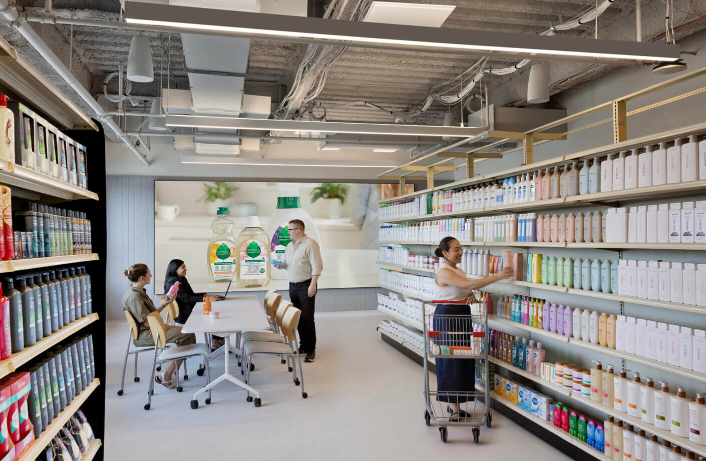 Interior view of a retail space with a woman organizing products on shelves, while two individuals discuss at a table in the background.