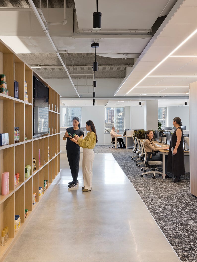 Two women engage in conversation near a shelving unit stocked with products in a sleek office space, featuring workstations in the background.