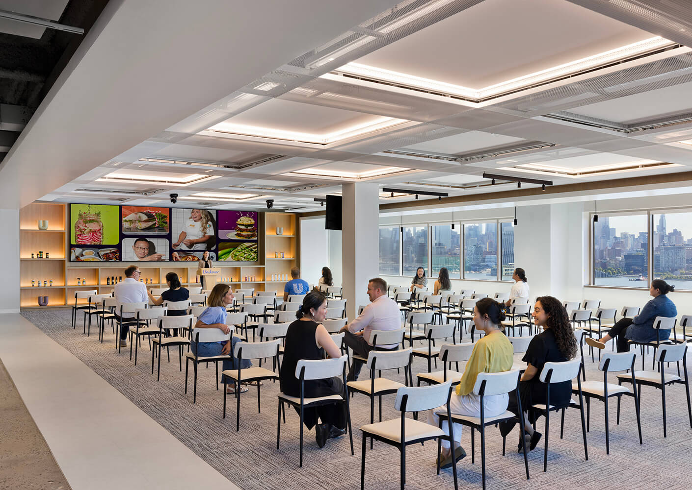 Spacious seminar room with rows of chairs, featuring a vibrant display of food photography and a city skyline view through large windows.