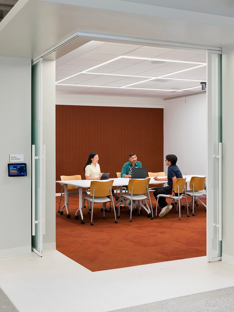 Four people engaged in a meeting around a table in a conference room with a warm-colored carpet and soft lighting.