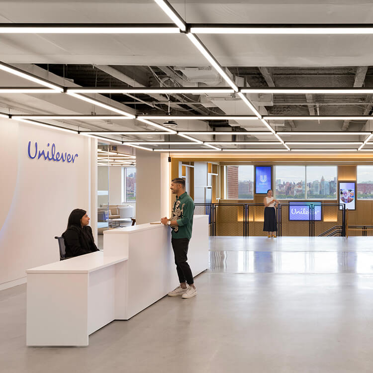 Reception area of Unilever office with two people interacting, modern lighting design, large windows, and branding displays in the background.