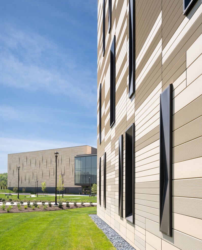 Exterior view of a building with textured siding, featuring angular windows and adjacent landscaping, under a clear blue sky.
