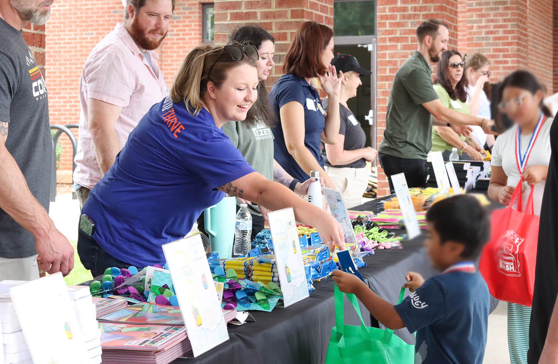 Community event with volunteers distributing supplies to children. Tables are filled with various educational materials and promotional items.