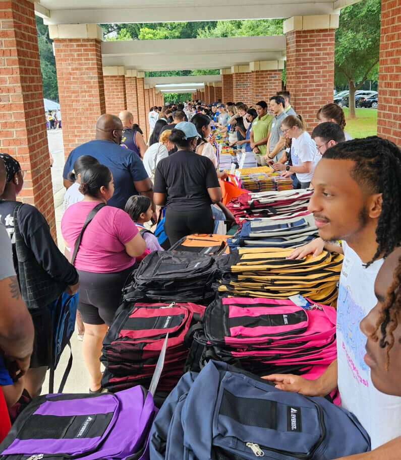 People line up to receive backpacks and school supplies at a community event, with tables showcasing various backpacks in different colors.