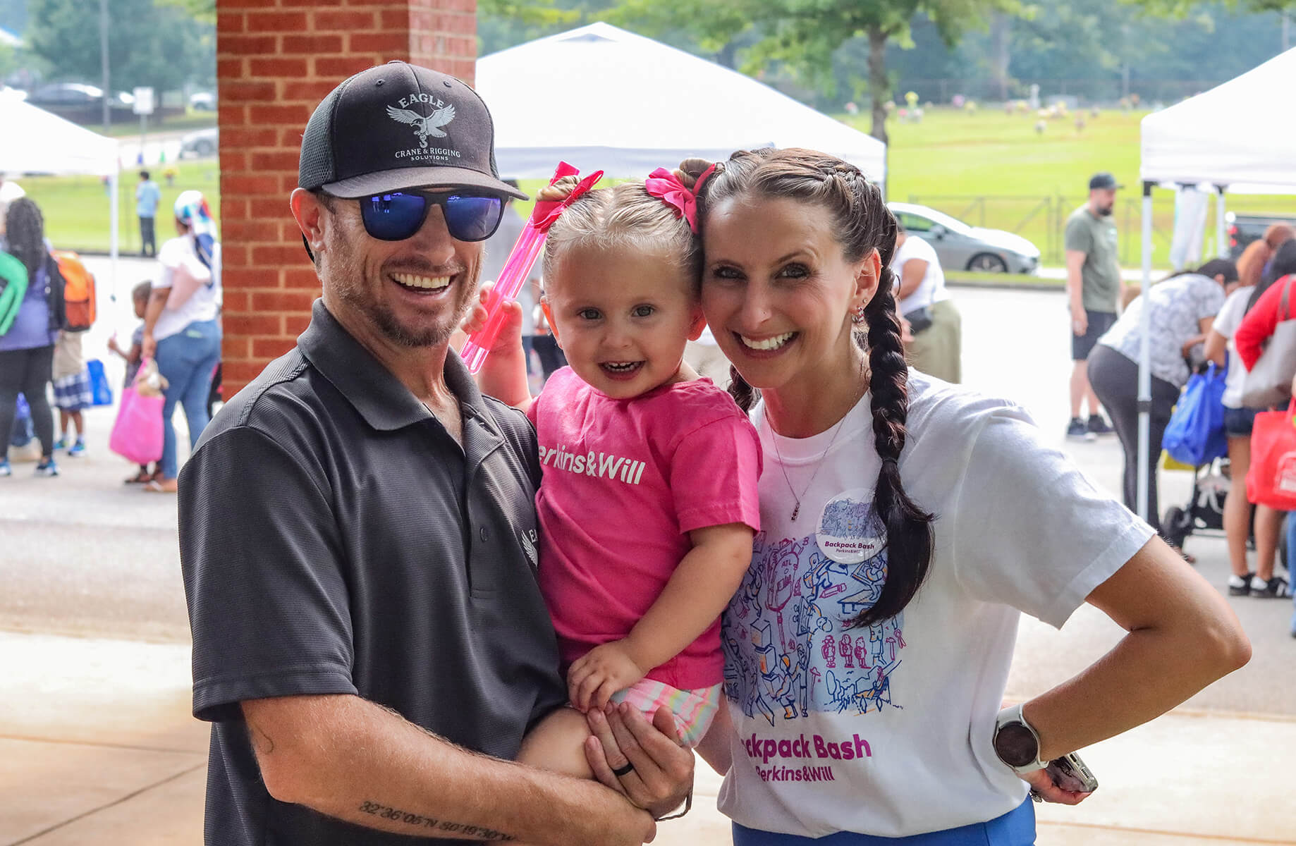 A man and woman pose with a young girl at a community event, all smiling. Tents and attendees are visible in the background.