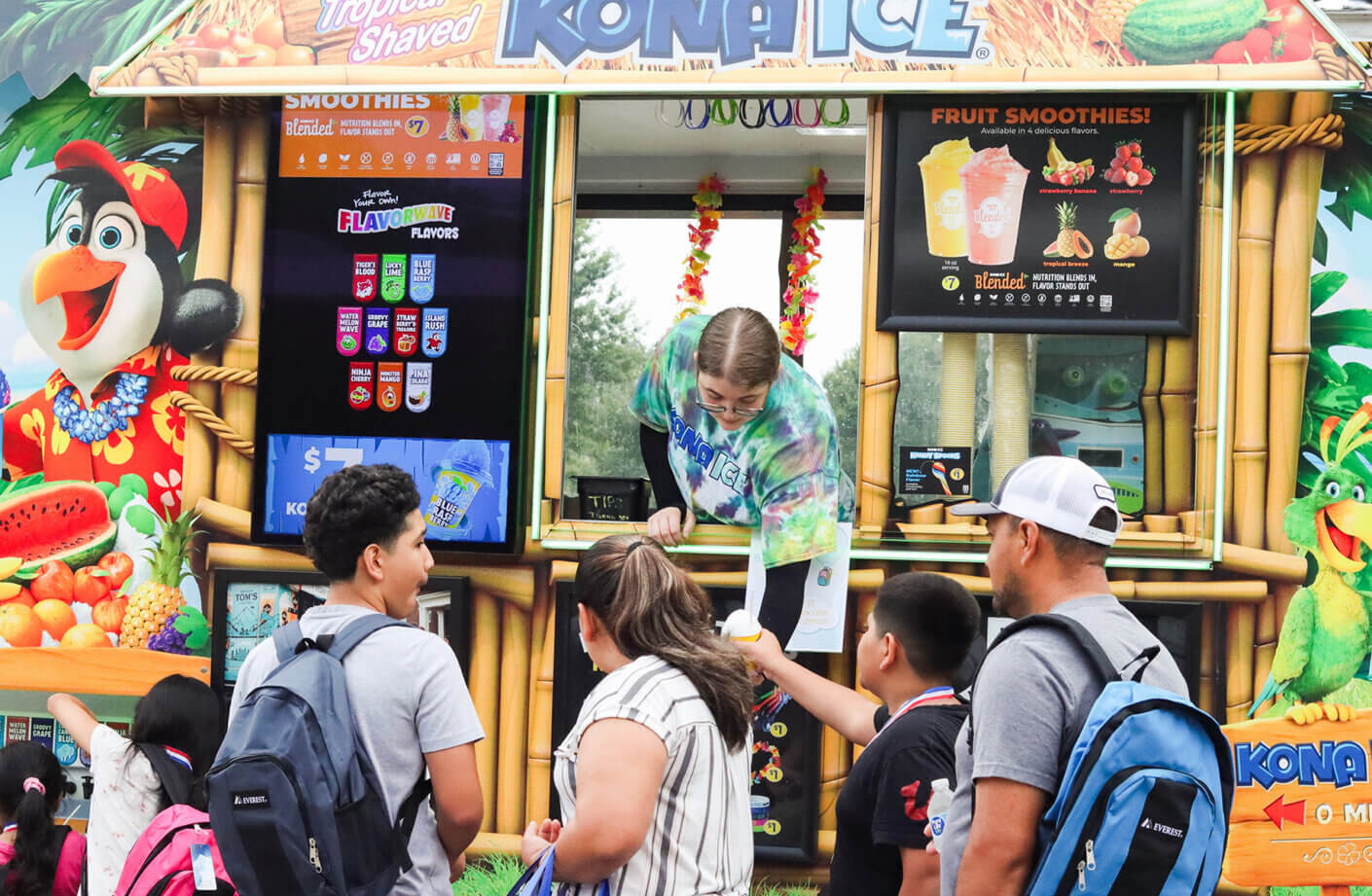 People lined up at a Kona Ice truck, ordering shaved ice and smoothies from a colorful menu. Staff member assisting customers at the window.