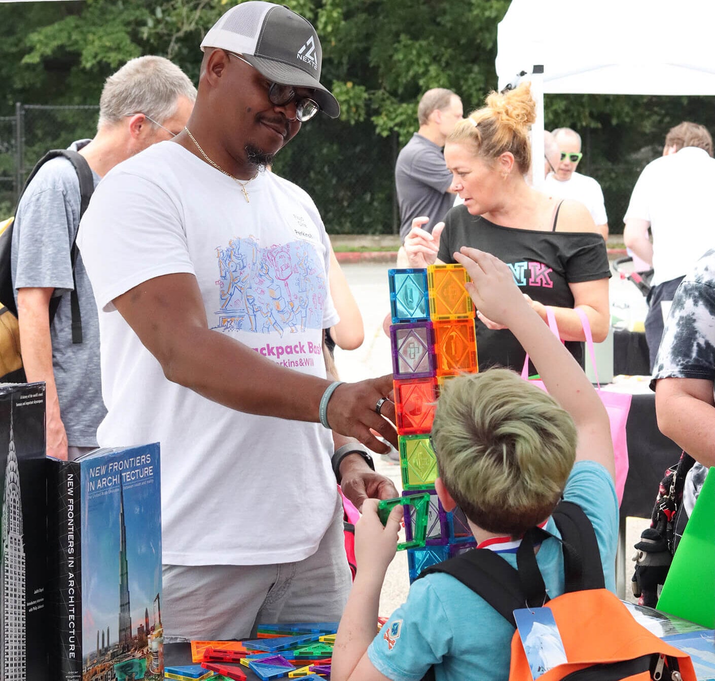 A man interacts with a child building a colorful tower at an outdoor event, surrounded by other attendees and a display of architectural books.