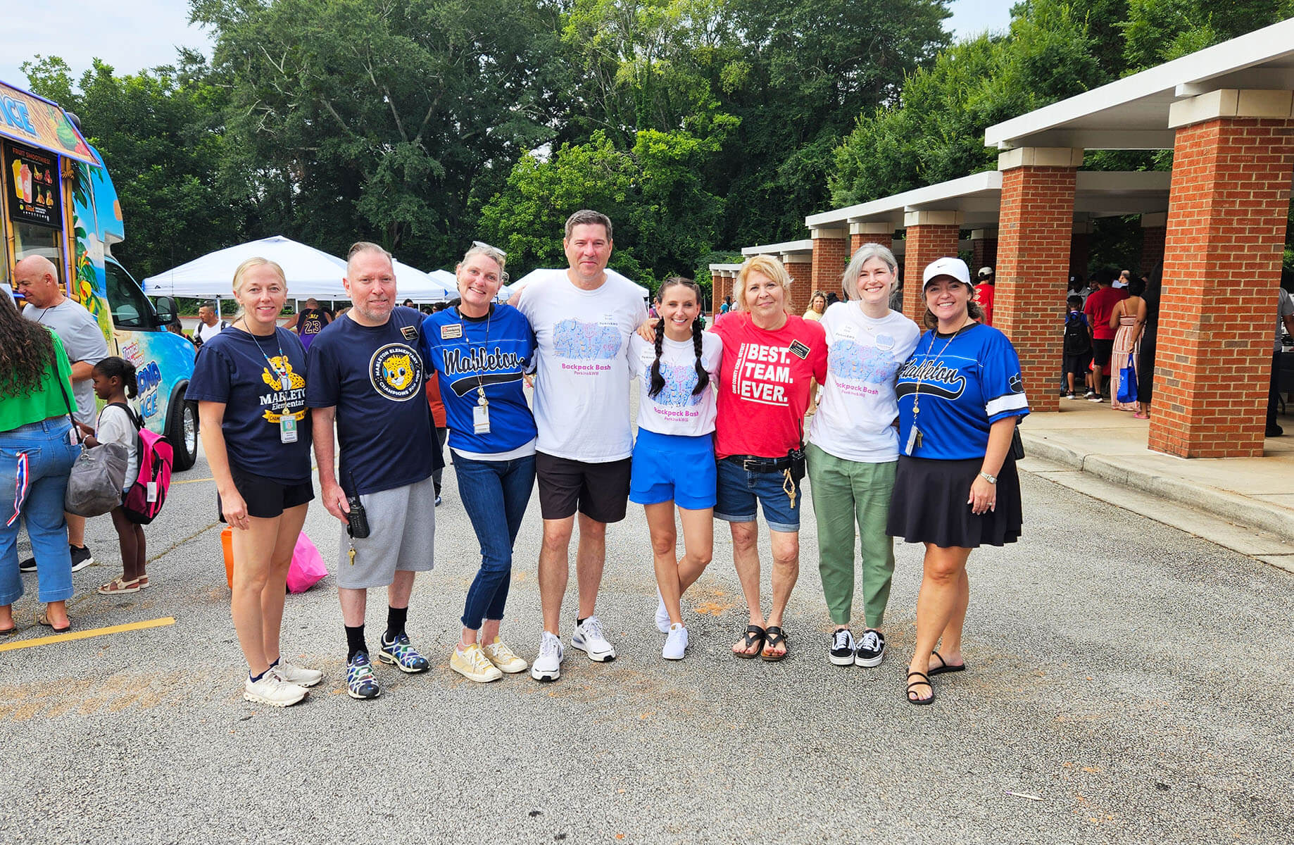 Group of eight people posing for a photo at an outdoor event, with food trucks and tents in the background, promoting community engagement.