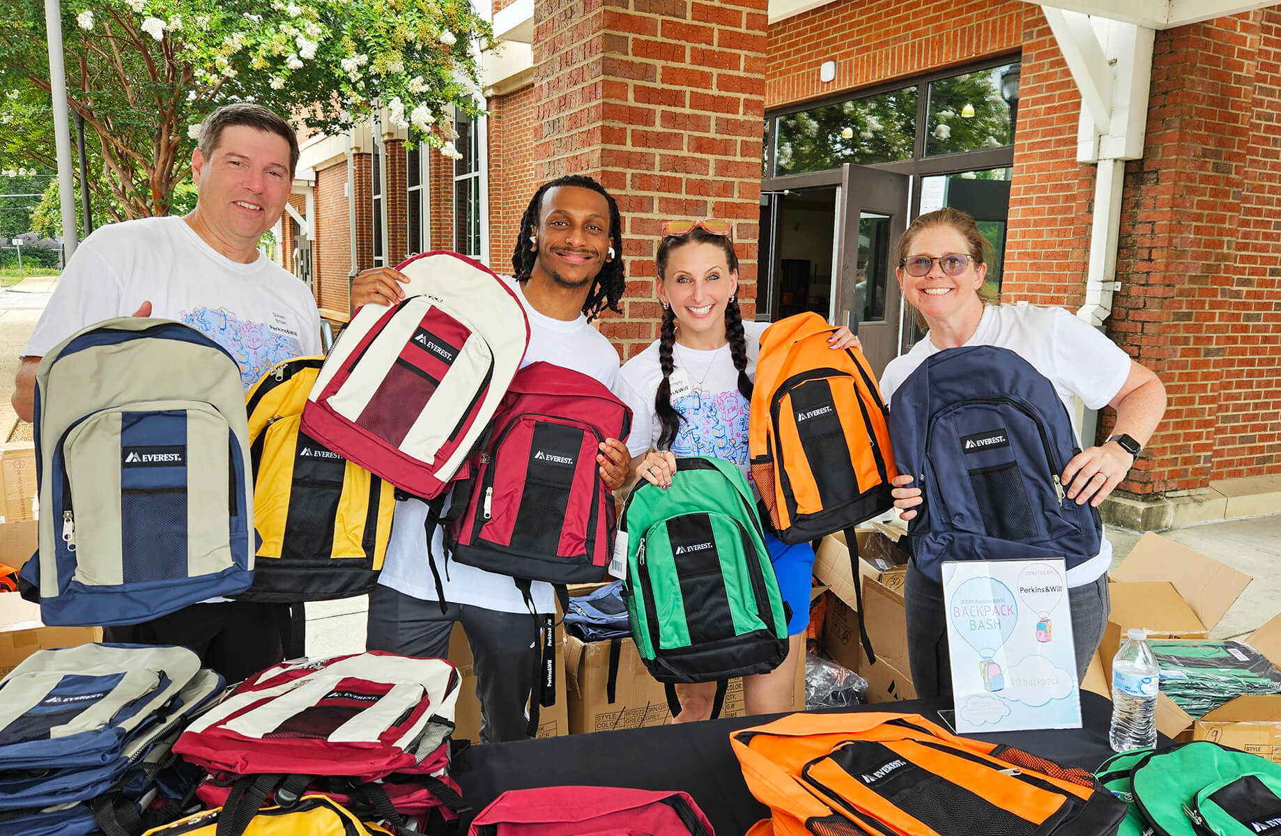 Four volunteers hold colorful backpacks at a community event. Boxes of additional backpacks are visible on a table.