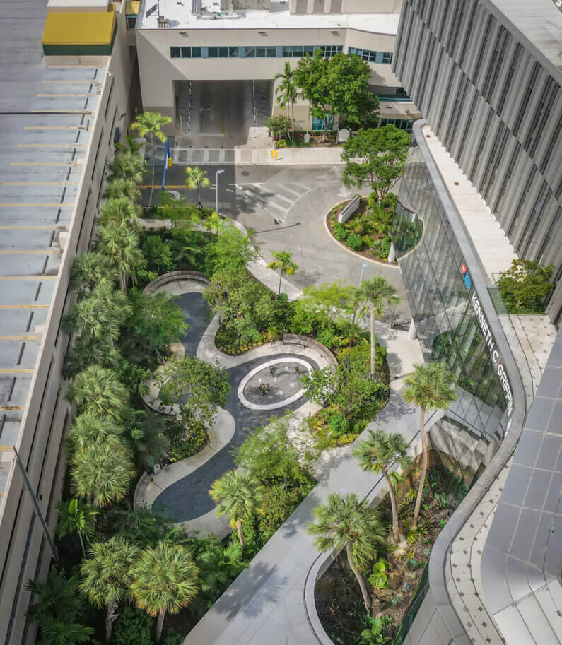 Aerial view of a landscaped area featuring winding pathways, greenery, and a glass building, situated in an urban environment.