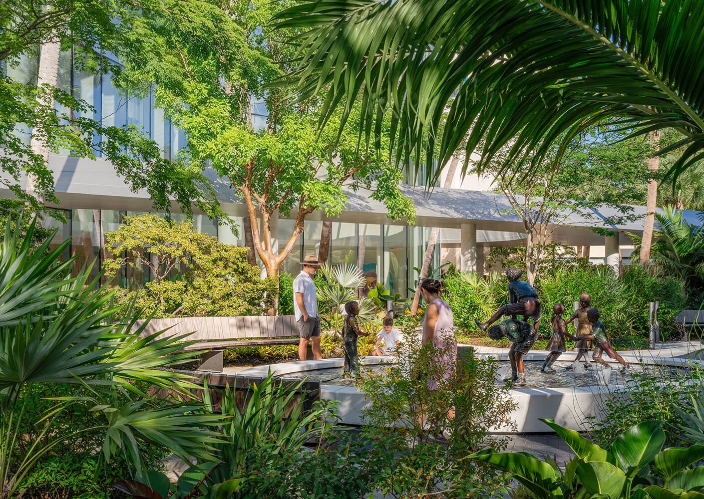 Visitors interact with sculptures and greenery in an outdoor space, featuring trees and modern architecture in the background.