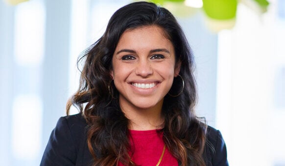 Smiling woman with long, wavy hair, wearing a black blazer and red blouse, stands in a bright office setting with greenery.