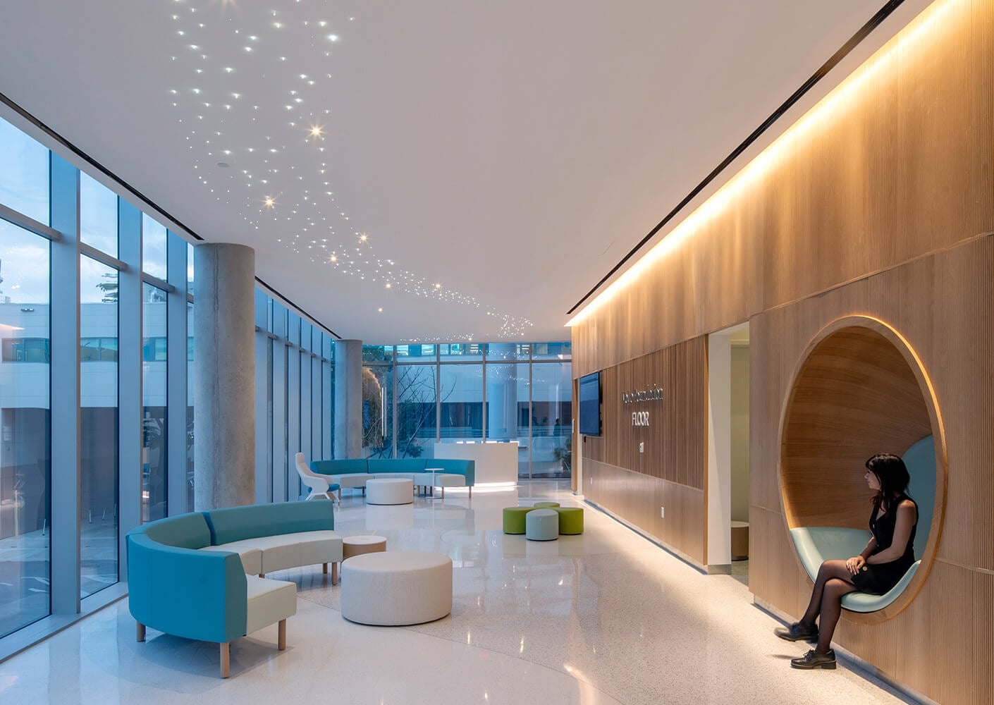 Interior of a reception area featuring large windows, unique seating, and ambient lighting, with a person sitting in a circular nook.