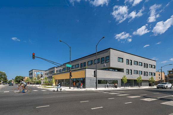 New urban building at a street corner with cyclists, pedestrians, and a clear blue sky. Features large windows and landscaped surroundings.