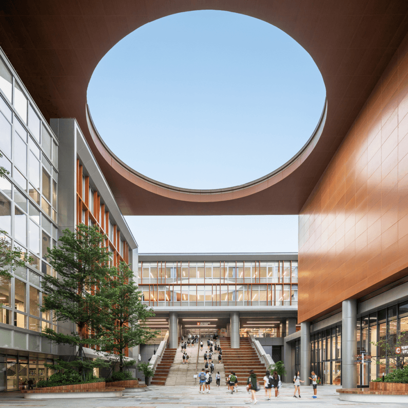 School entrance with a circular opening above, surrounded by buildings and large staircases, with students walking through the space.