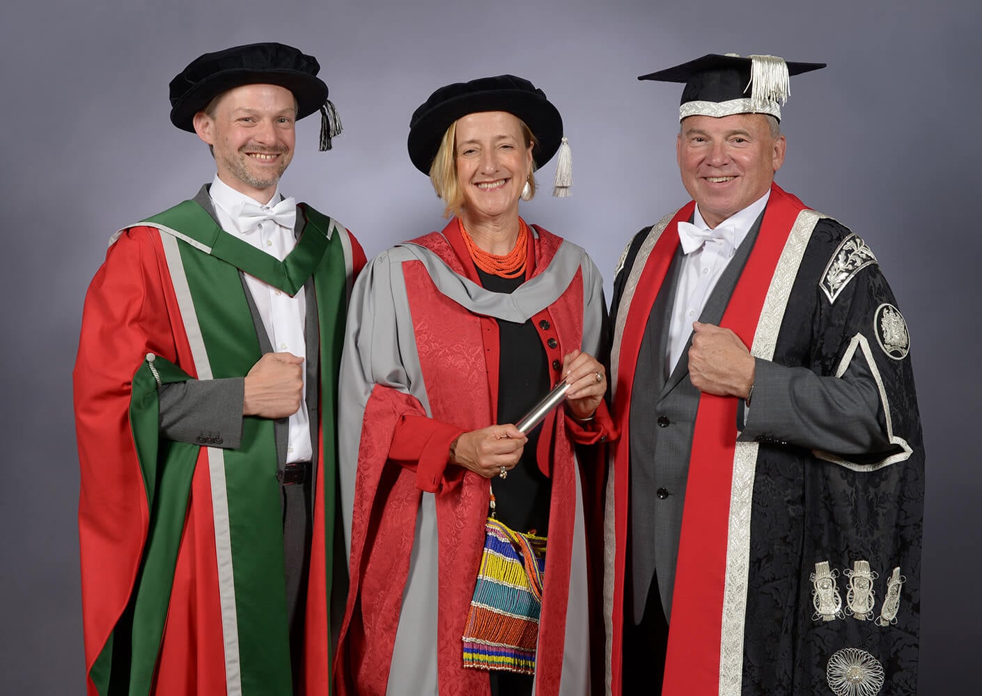Three individuals in academic regalia pose for a photograph, showcasing their ceremonial attire and accessories against a gray backdrop.