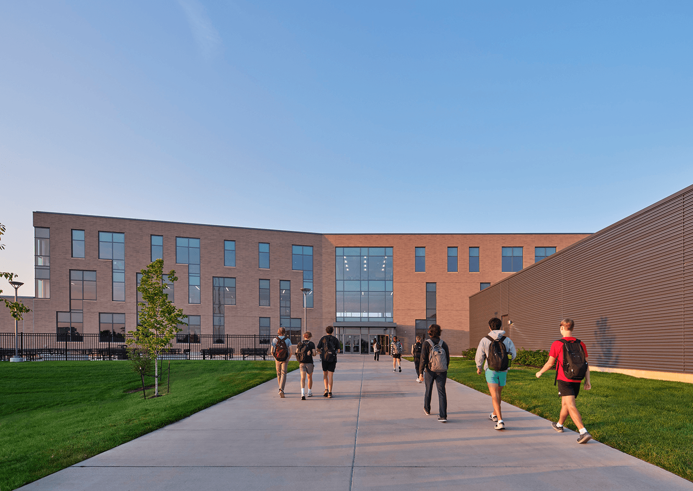 Students walk along a path towards a large school building with multiple windows and green landscaping. Clear sky in the background.