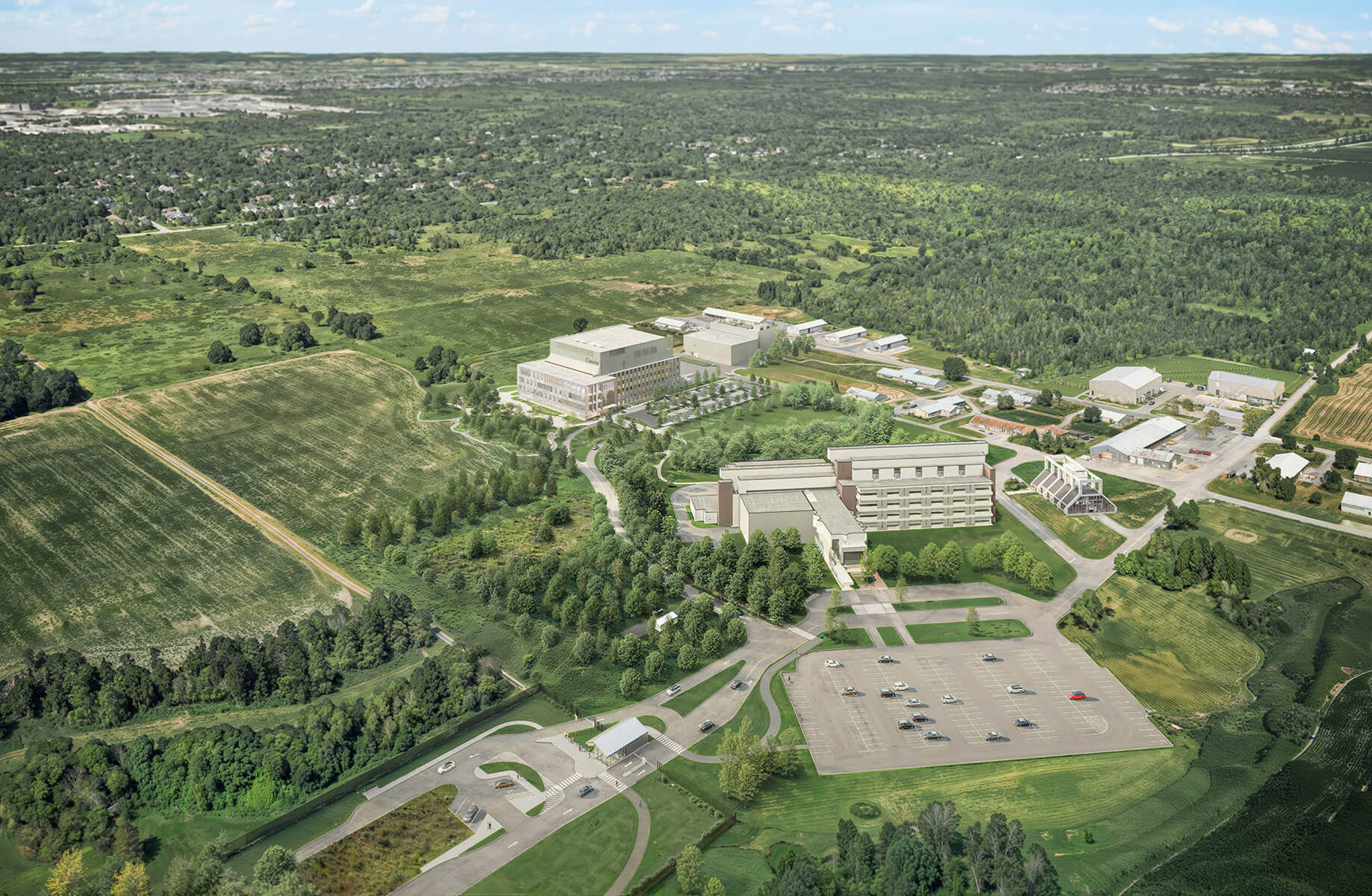 Aerial view of a large facility surrounded by green fields, trees, and parking areas, situated in a landscape with distant buildings and hills.