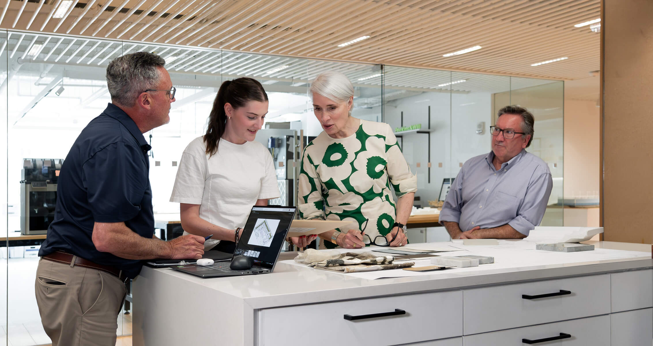 Robin Osler, workplace practice leader for the Boston studio collaborating with colleagues discussing design plans at a workspace, with a laptop and samples on the table. Collaborative atmosphere.