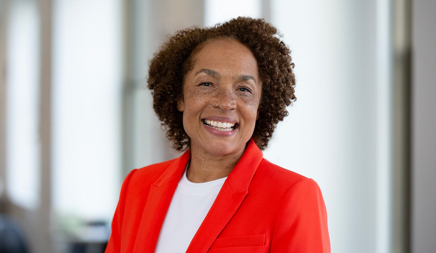 Professional woman with curly hair wearing an orange blazer and a white shirt, smiling in a bright indoor setting.