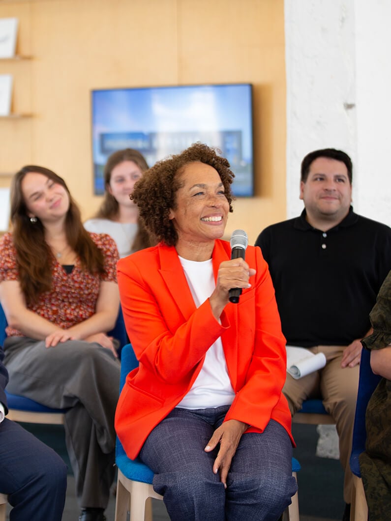 A woman in an orange blazer speaks into a microphone during a panel discussion with engaged audience members in a bright, open space.