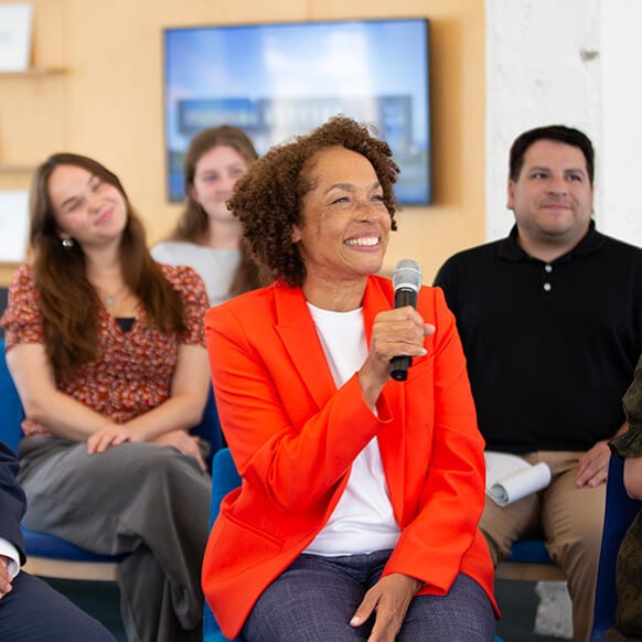 Group discussion featuring a woman in an orange blazer speaking into a microphone, with an audience attentively listening in a bright, open space. 